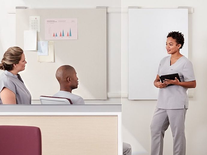 Three people in scrubs conversing behind a nurses station in front of a tackboard and a whiteboard.