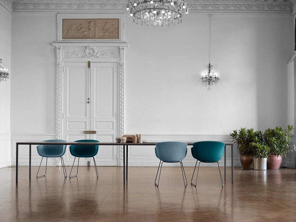 A long wood conference table surrounded by upholstered grey and blue guest chairs in a room with a wood floor and glass chandelier.