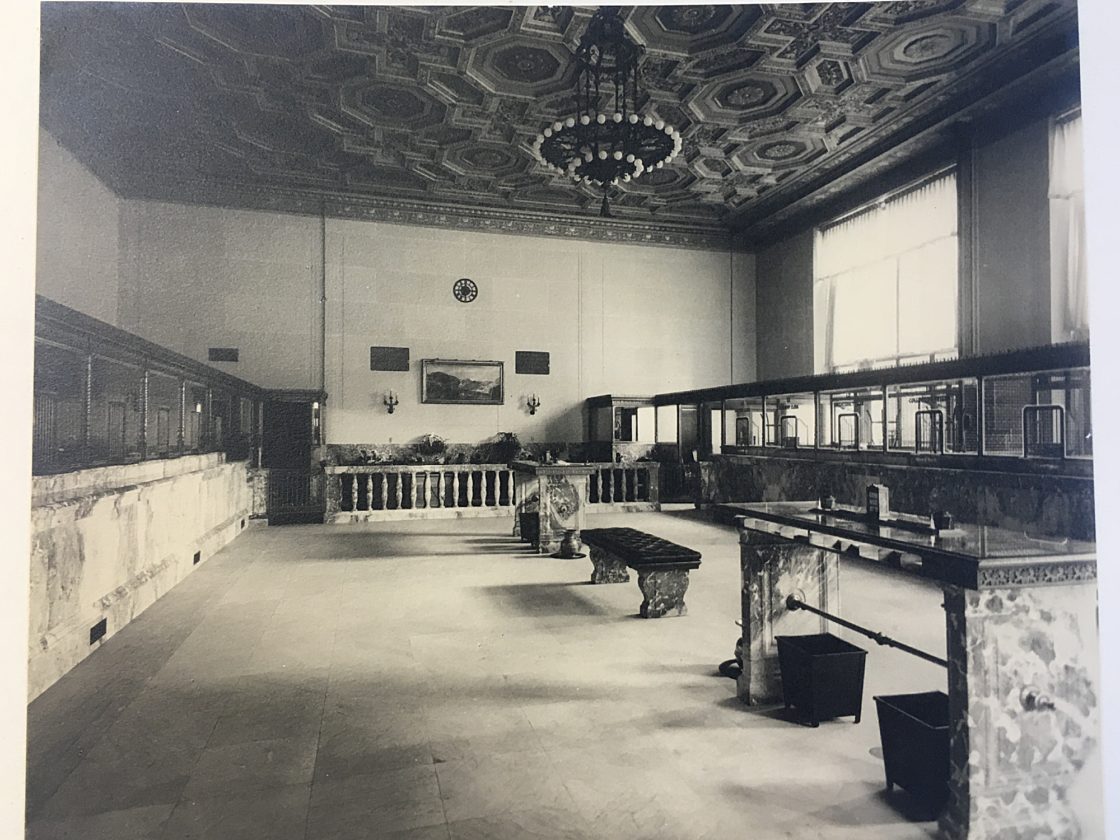 A black and white historical photo of the interior of our current headquarters building when it was a bank with the same marble floors and stucco ceiling.