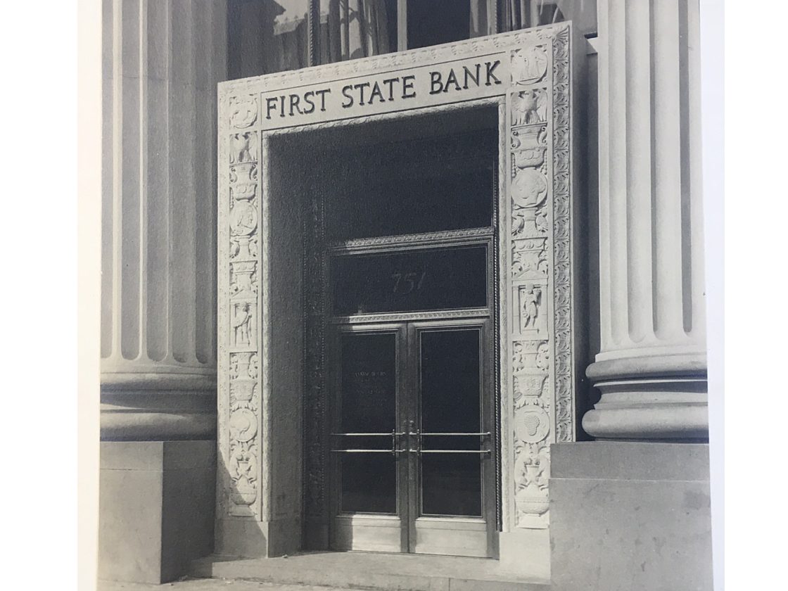 A black and white photo of the MarxModa headquarters showcasing the carved front facade with First State Bank chiseled in stone.