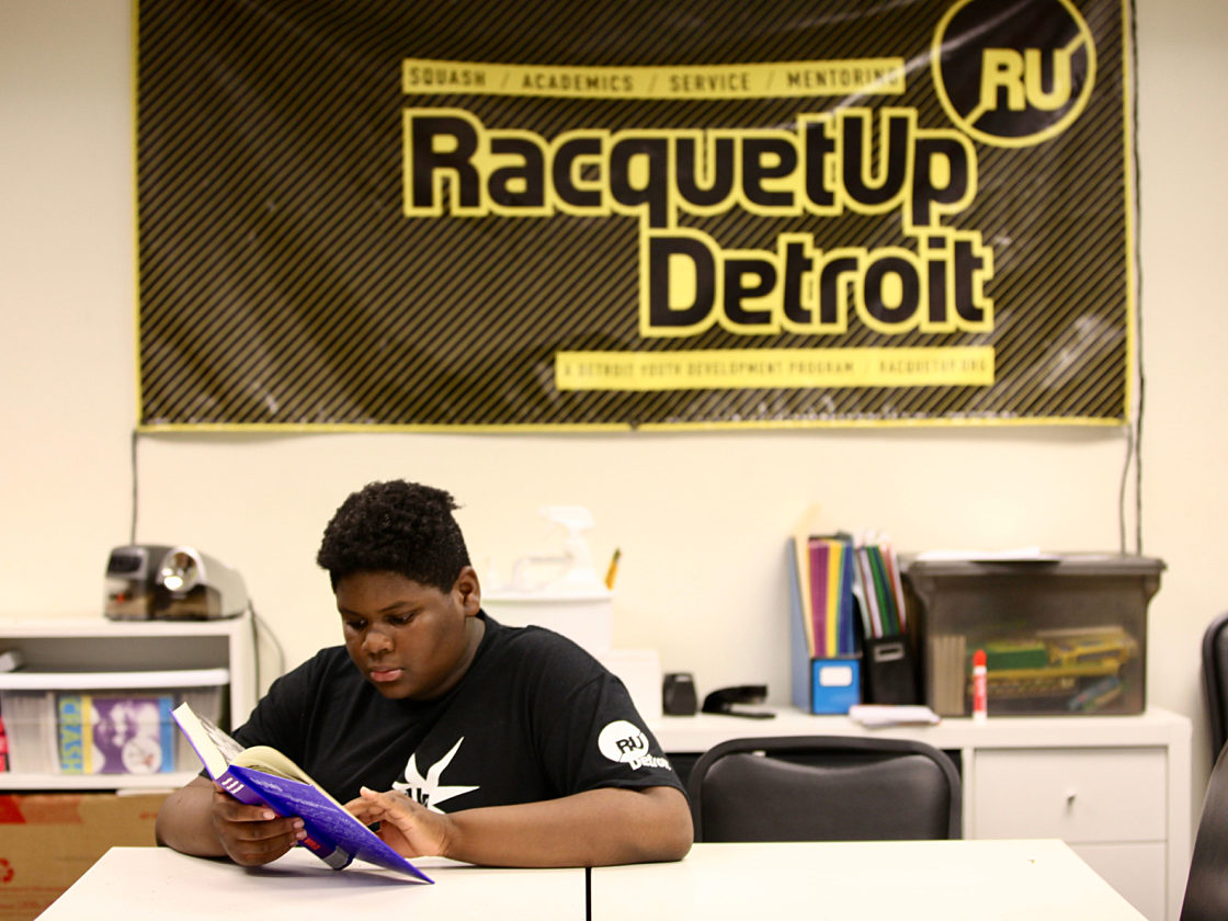 An image of a boy in a Racquet Up t-shirt reading a book at a desk in front of a Racquet Up banner.