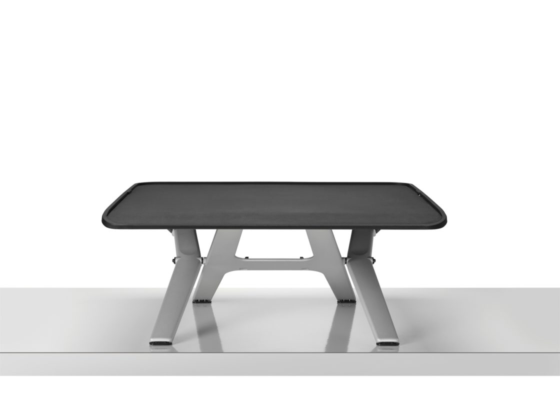 A Monto Sit-to-Stand Riser with a black top and silver legs on a silver surface in front of a white wall.
