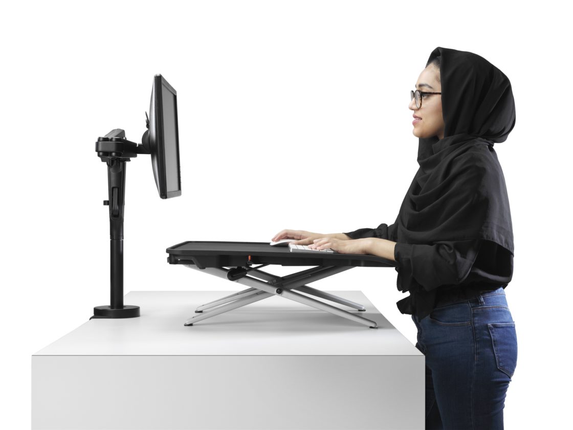 A woman standing at a white desk working on a keyboard set on top of a Monto Sit-Stand Desk looking at a screen mounted at eye level on an Ollin Arm.