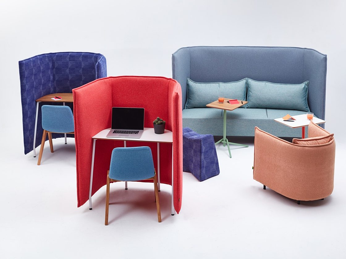 A Cloud Desk with a high pink fabric desk in front of a light blue Cloud Sofa with a blue high-back surround next to a peach Cloud Chair.