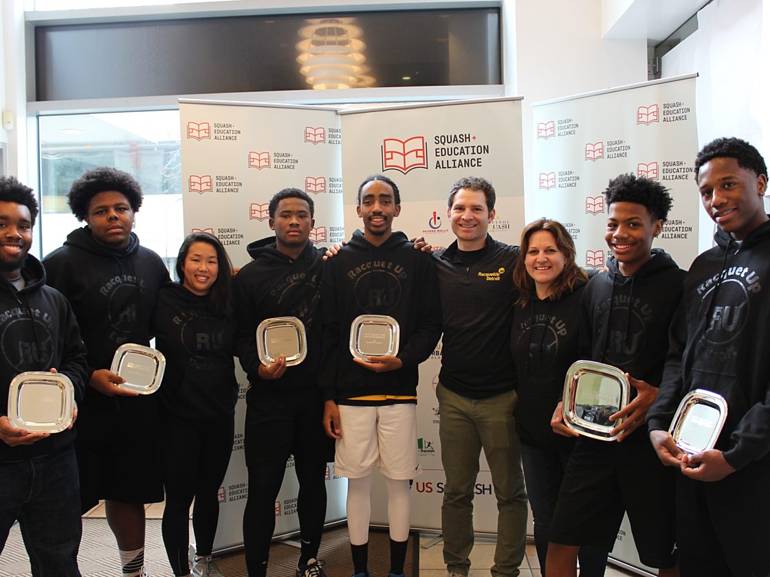 A group of kids and coaches in Racquet Up sweatshirts holding silver award plaques in front of a Squash Education Alliance banner.