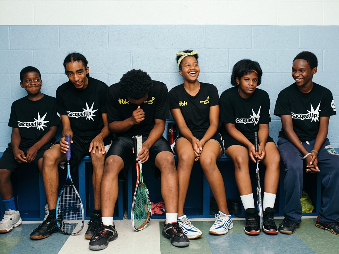 A group of kids in Raquet Up t-shirts and athletic clothing with squash racquets.