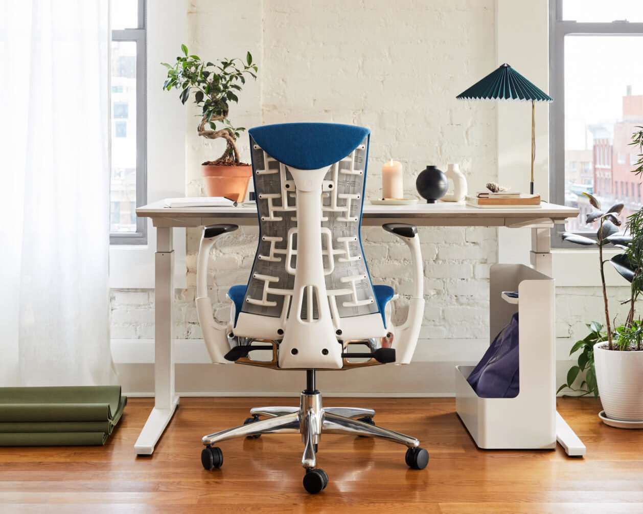 The back of an Embody chair with a white frame and blue upholstery in front of a sit-to-stand desk with Ubi Storage and a HAY table lamp in a home office.
