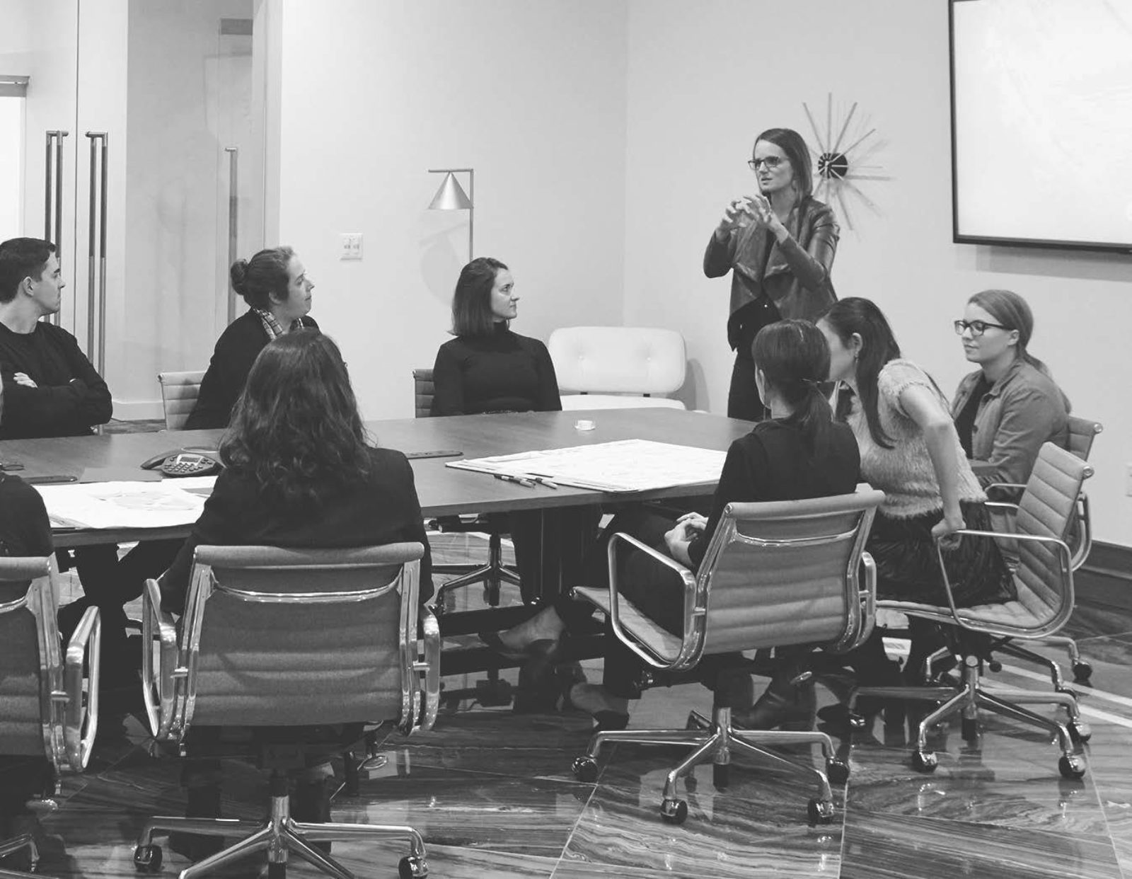 A group of designers in the MarxModa family room sitting at the conference table watching a woman stand and present.