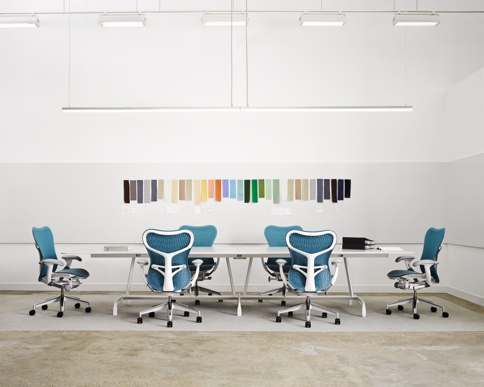 A conference room with a long white table surrounded by blue and white Mirra 2 chairs with a colorful art installation on the wall behind the table.