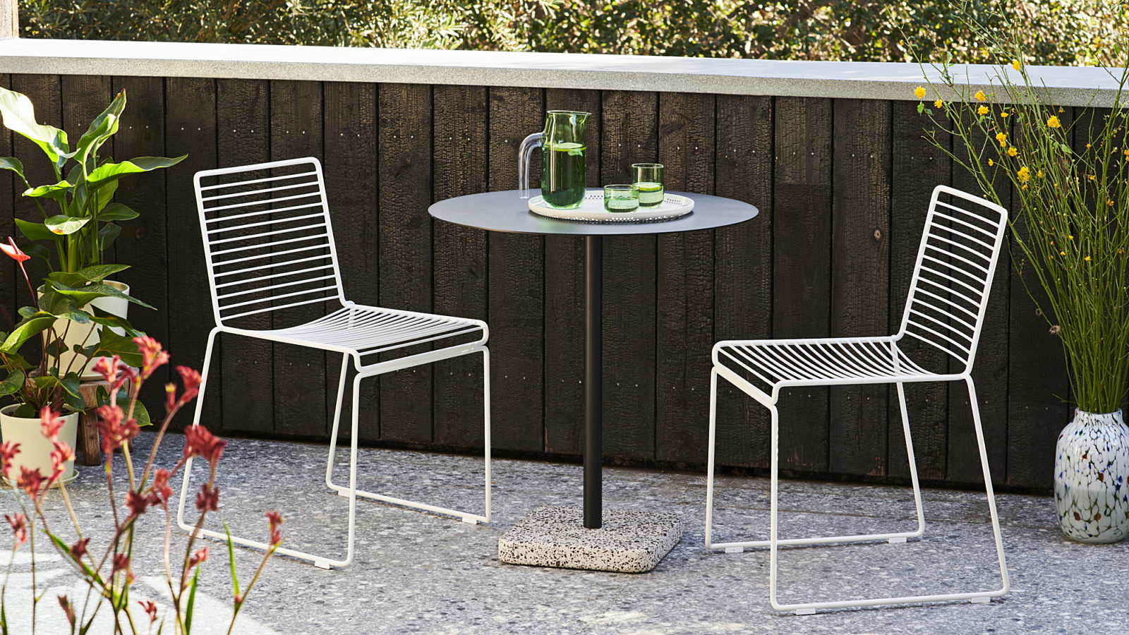 Two white HAY Hee Dining Chairs facing each other across a black round HAY Terrazzo Table on a stone patio outside.