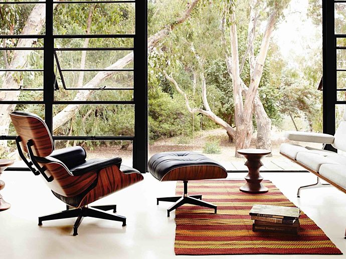A white Eames Sofa and black Eames Lounge and Ottoman in a light-filled living room with two Eames Walnut Stools.