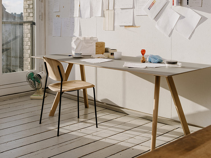 A home office with a wood HAY Soft Edge 10 chair with a black frame in front of a wood Pyramid Table filled with loose papers and office supplies.