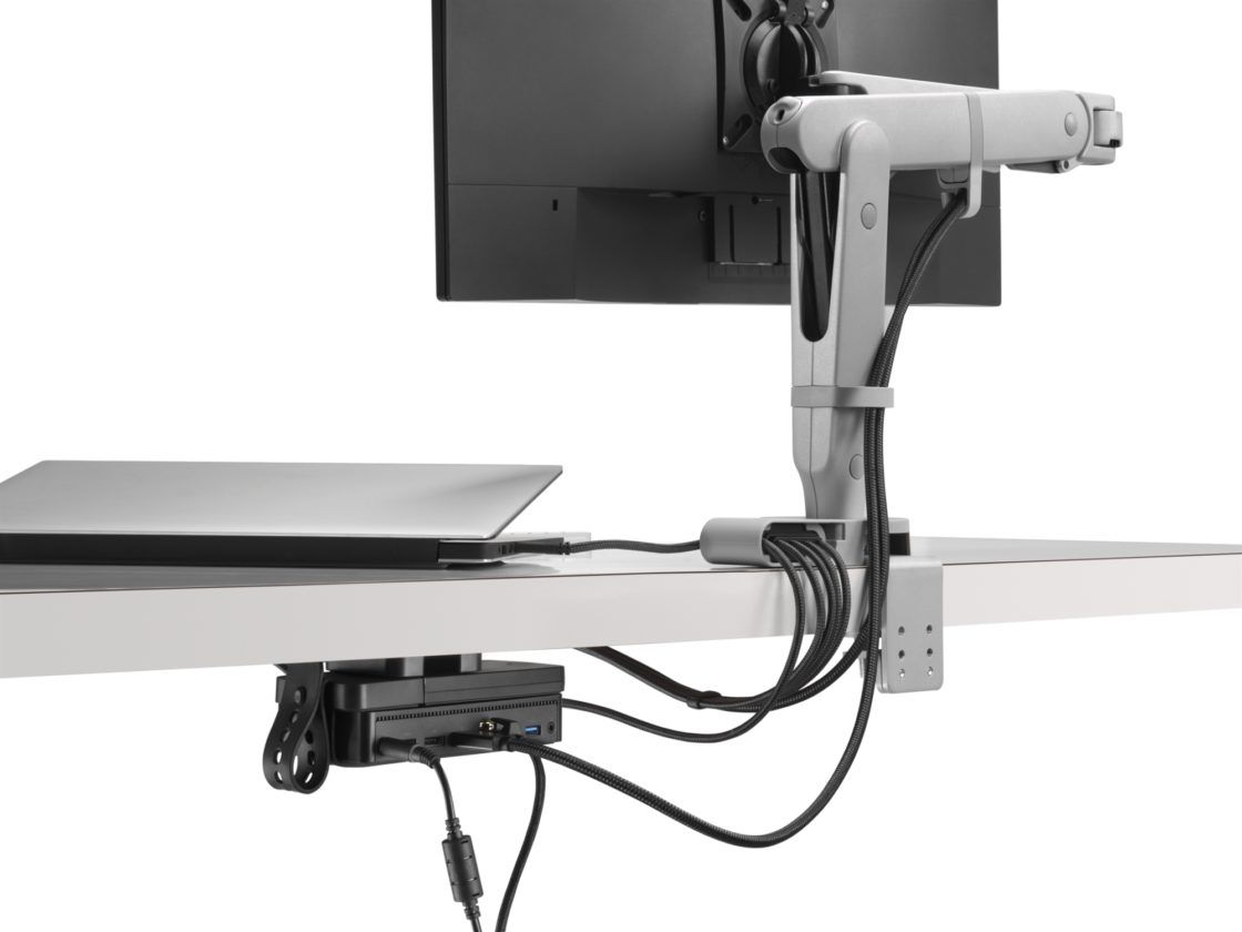 A back view of a Loop Micro Mount attached to the bottom of a worksurface holding a device that is attached to a laptop sitting below an Ollin Arm.