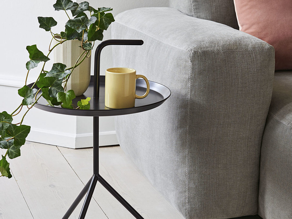 A black HAY Don't Leave Me Side Table holding books and a coffee mug next to a sofa.