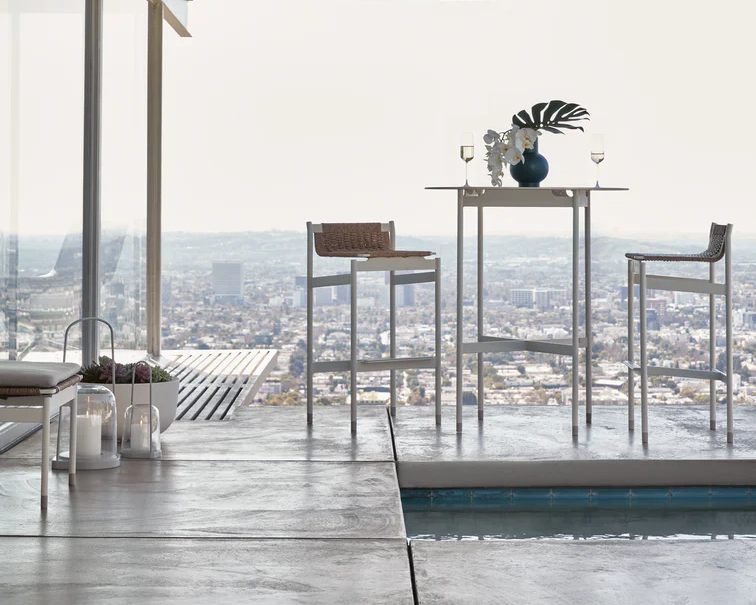 Two white Sommer Barstool facing eachother across a white metal table sitting on a deck overlooking a large cityscape.
